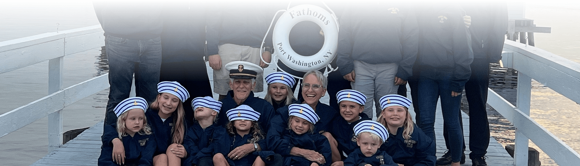 A group of adults and children on a dock holding a life preserver that says Fathoms, Port Washington, NY
