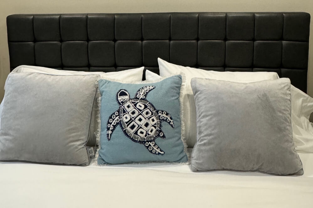 A vignette of pillows on a bed with white grey and blue pillows, the blue one with a sea turtle embroidered on it.