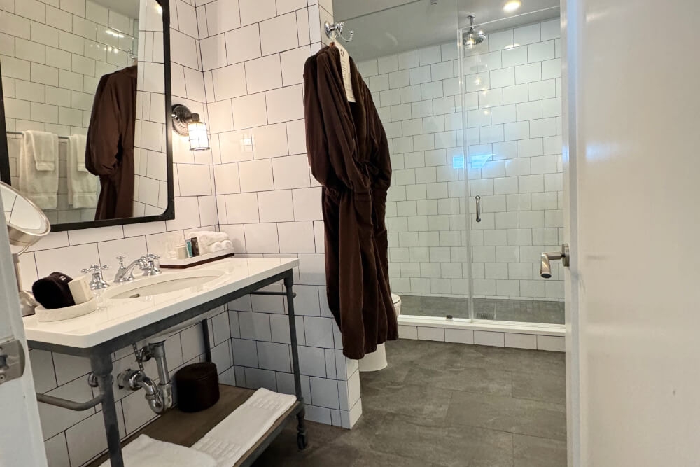 A bathroom with a shower with clear glass doors, a vanity with sink and mirror above it, and a brown robe hanging near the shower.