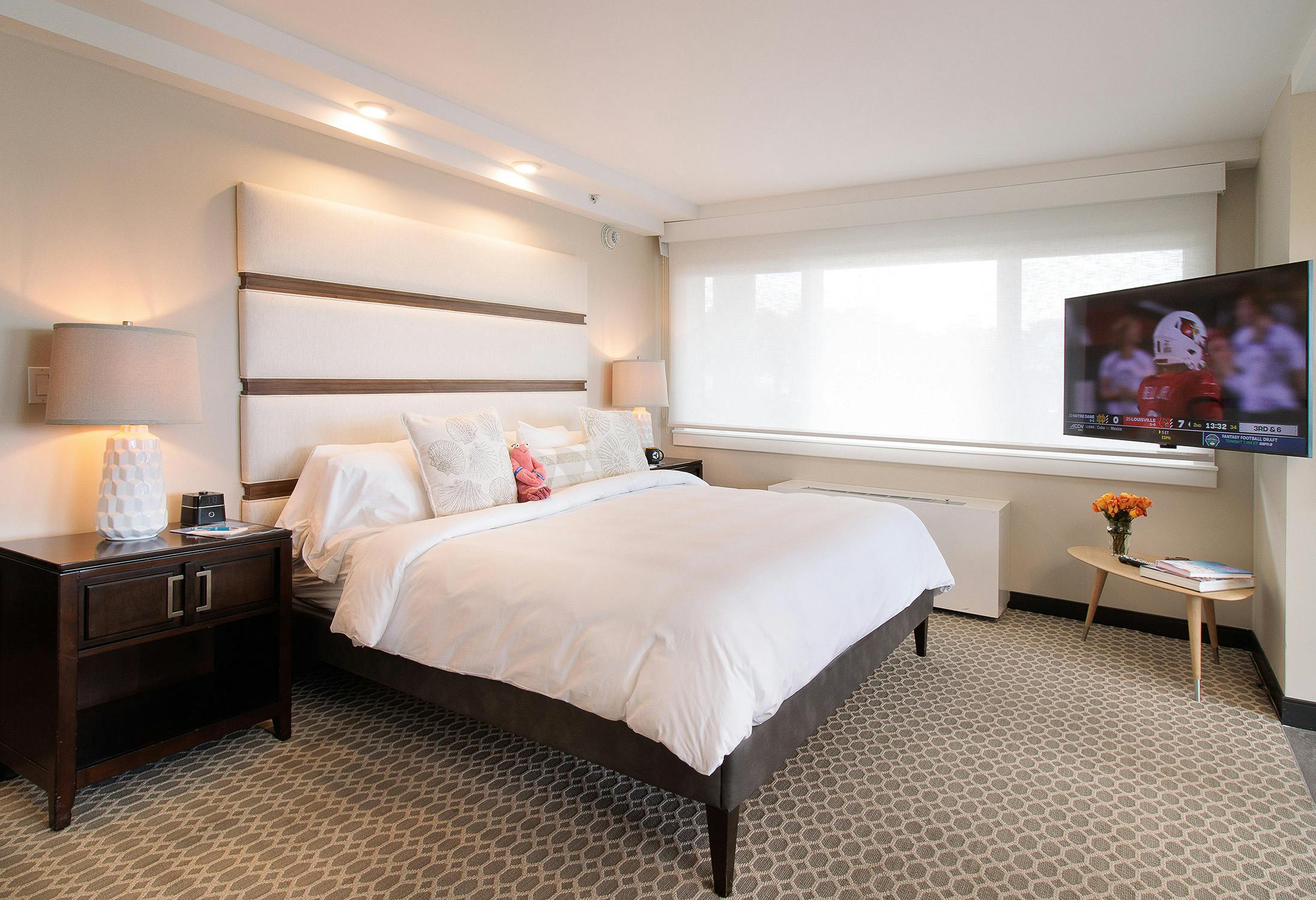 A bedroom with a bed with white bedding, wood nightstands on either side of the bed with lamps, and an alcove area with a mini fridge and coffee center.