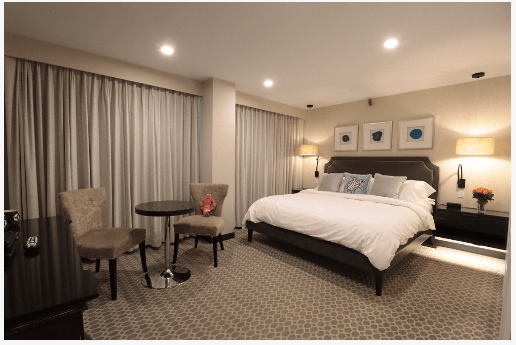 A bedroom with carpeted floor, a king bed with white bedding, night stands with lamps on either side of the bed, and a sitting area with a plush chairs and a small table.