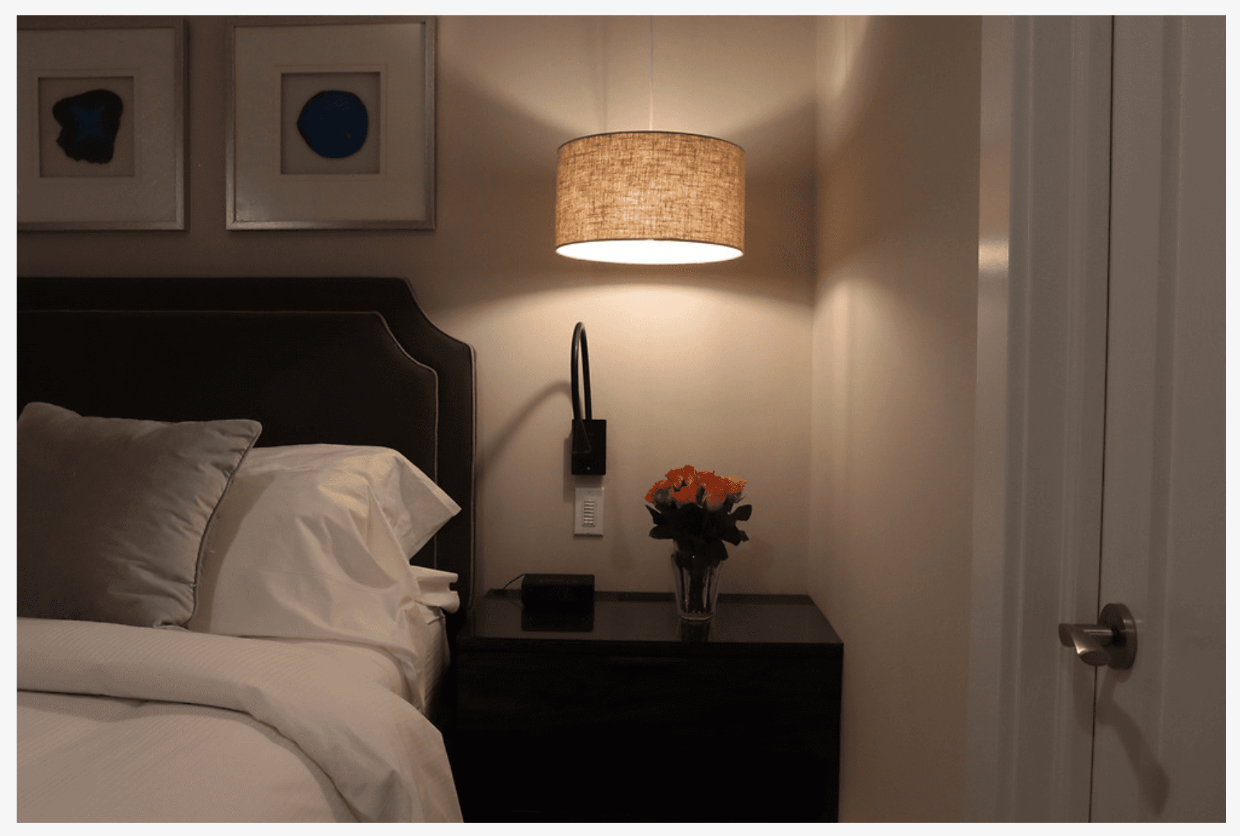A night table next to a bed with an overhead hanging fixture, reading light, and flowers.