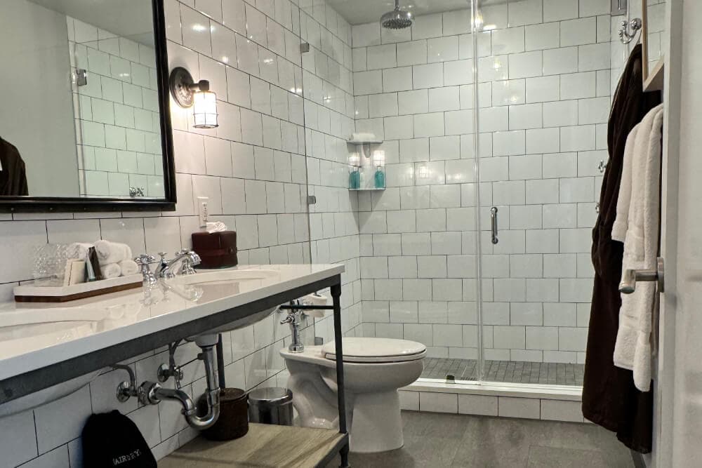 A bathroom with a double sink vanity, a mirror above the vanity, a shower with clear glass shower doors, a toilet, white towels, and a brown robe.