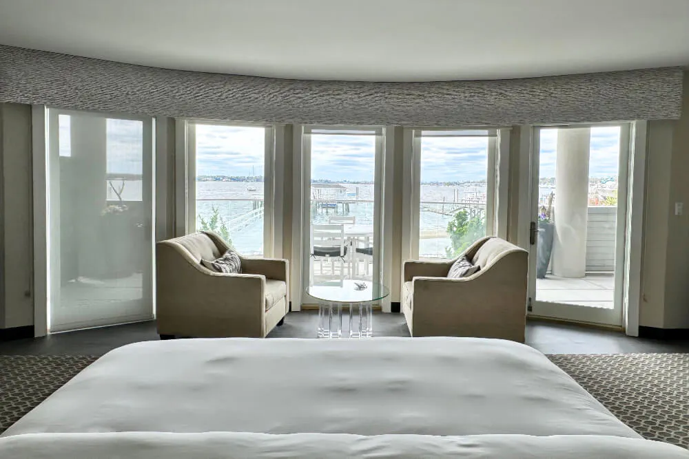 A bedroom with a bed, sofa, and sitting area with 2 chairs and table in front of a bank of windows overlooking a marina.