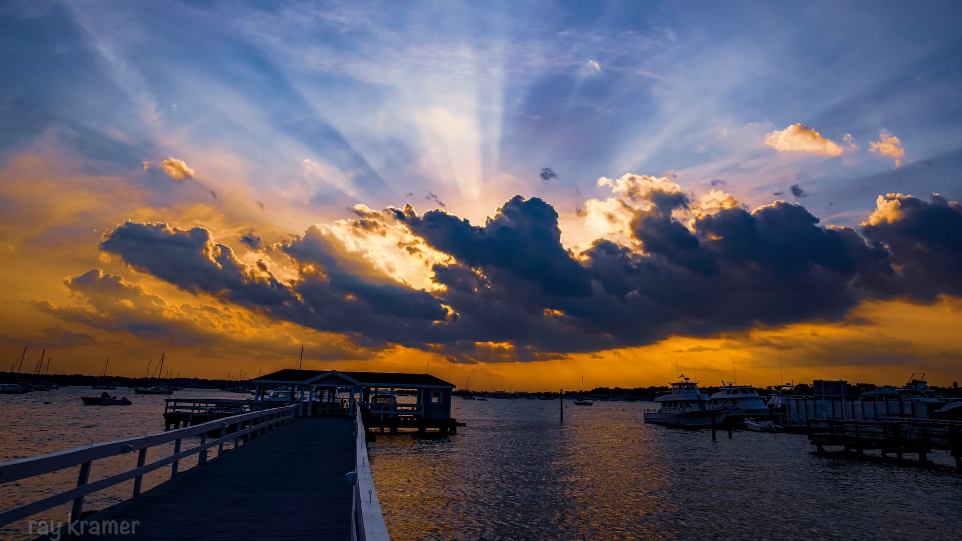 A wooden pier stretches into a tranquil body of water under a dramatic sunset with vibrant clouds and rays of light.