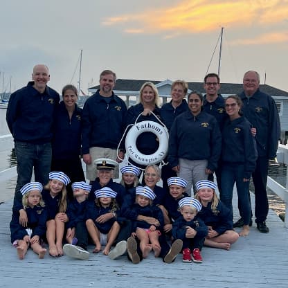 A group of adults and children on a dock holding a life preserver that says Fathoms, Port Washington, NY